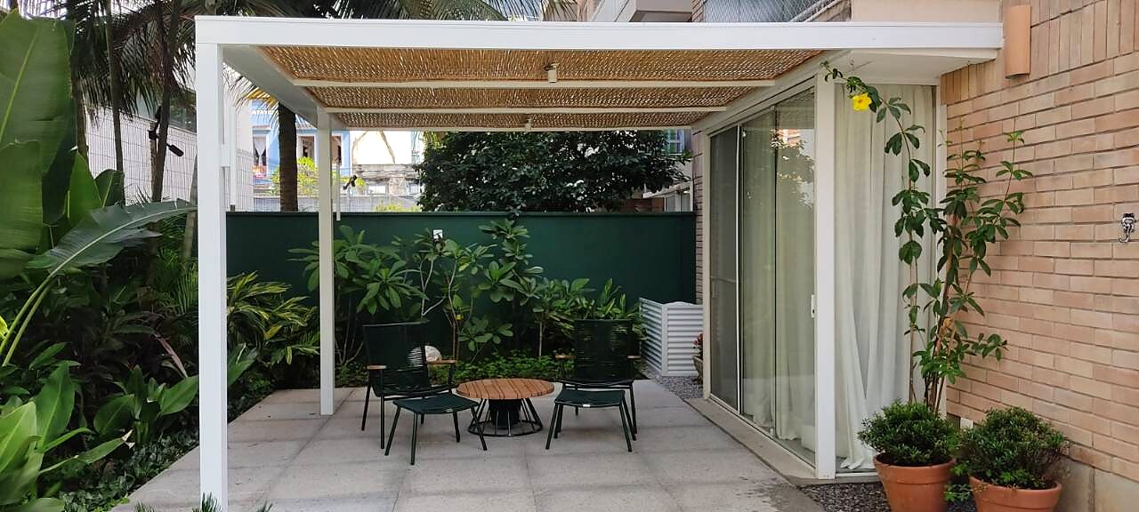 Pergolado em alumínio branco com fechamento superior em bambu sobre área externa térrea com jardim tropical